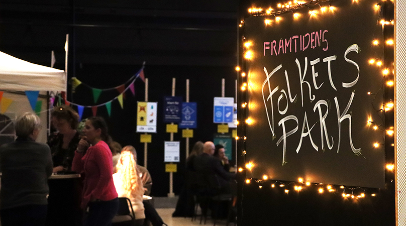 På en dörr står en skylt med texten "Framtidens Folkets park". Inne i rummet syns människor och volanger.