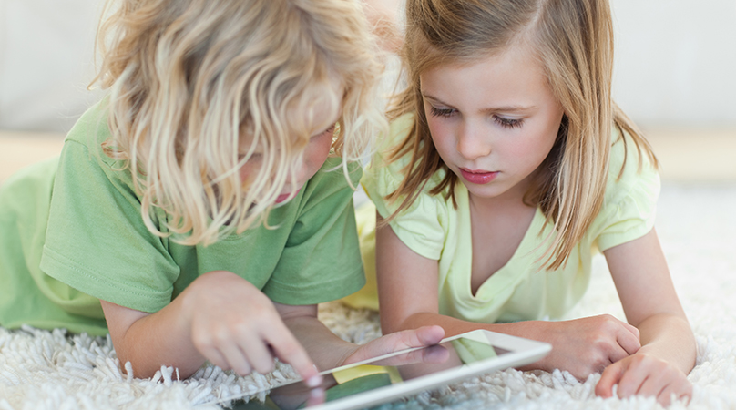Två barn ligger på golvet och kollar på en iPad.