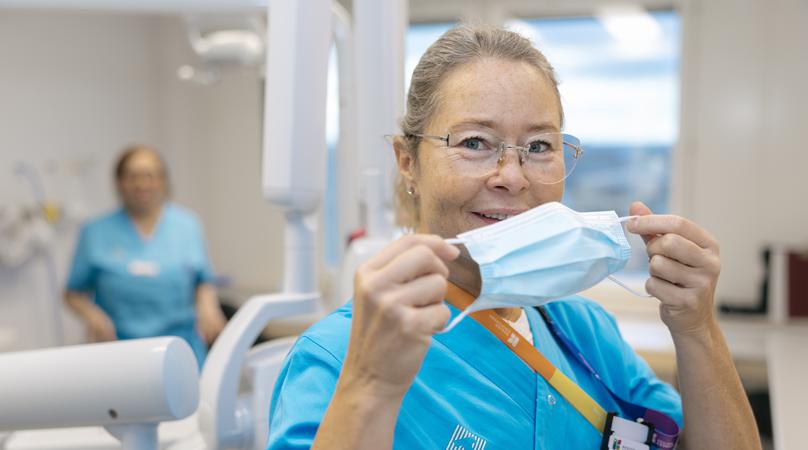 En kvinna håller ett munskydd framför ansiktet, bakom henne syns en tandläkarstol.