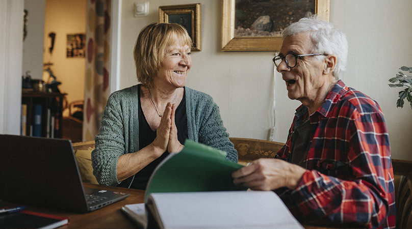 Äldre kvinna och man sitter vid bord och samtalar. 