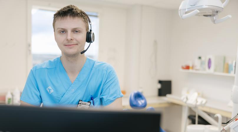 En man med headset står vid en dator, bakom honom syns en tandläkarstol.