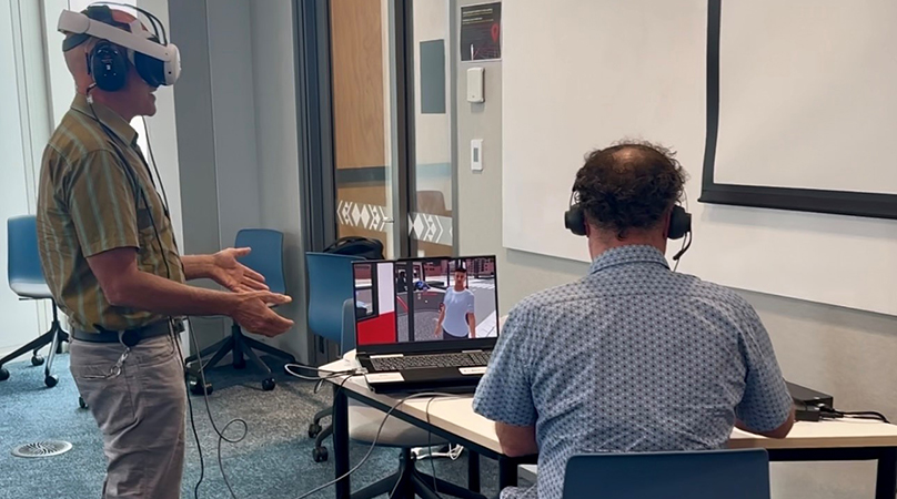En man har ett VR-headset på sig och en annan man sitter vid en dator som speglar vad mannen med VR ser. 
