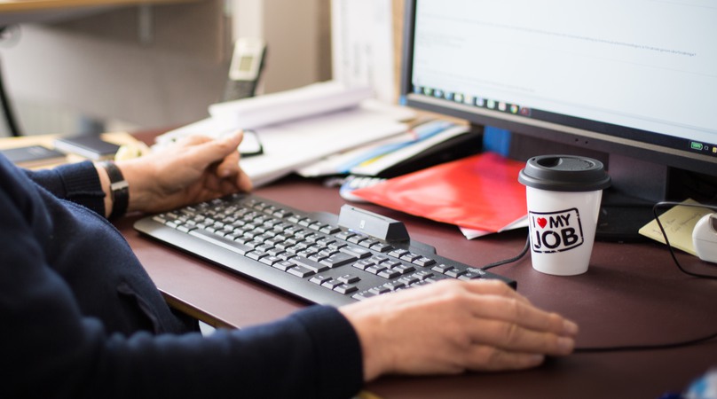 En person sitter vid ett skrivbord med en datorskärm. På skrivbordet st¨år även en kaffemugg där det står "i love my job".