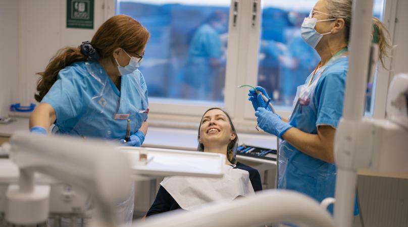 Två kvinnor i vårdkläder står bredvid en tandläkarstol med en patient i.