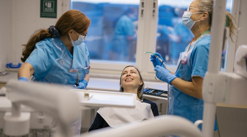 Två kvinnor i vårdkläder står bredvid en tandläkarstol med en patient i.