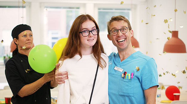 Till höger står en vårdklädd man och håller om en yngre kvinna, bakom dom står en kvinna med ballonger och i rummet yr guldiga stjärnor.