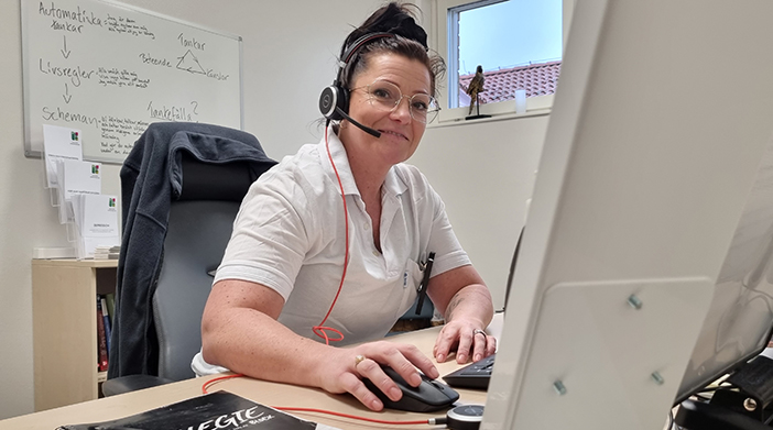 En kvinna sitter vid ett skrivbord. På huvudet har kvinnan ett headset och i handen en datamus.