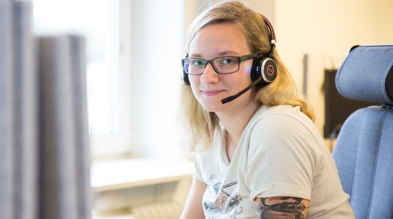 En kvinna sitter i en kontorsstol med headset och ler mot kameran.