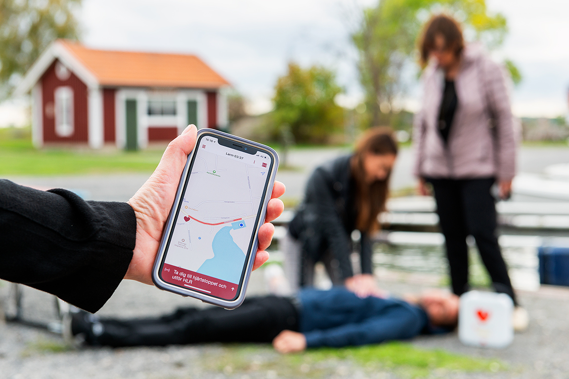Bilden illustrerar ett misstänkt hjärtstopp. Två personer hjälper en livlös människa, dne ena utför HLR och jämte dem ligger en hjärtstartare. I förgrunden håller en hand upp en mobiltelefon som har appen Sms-livräddare uppe.