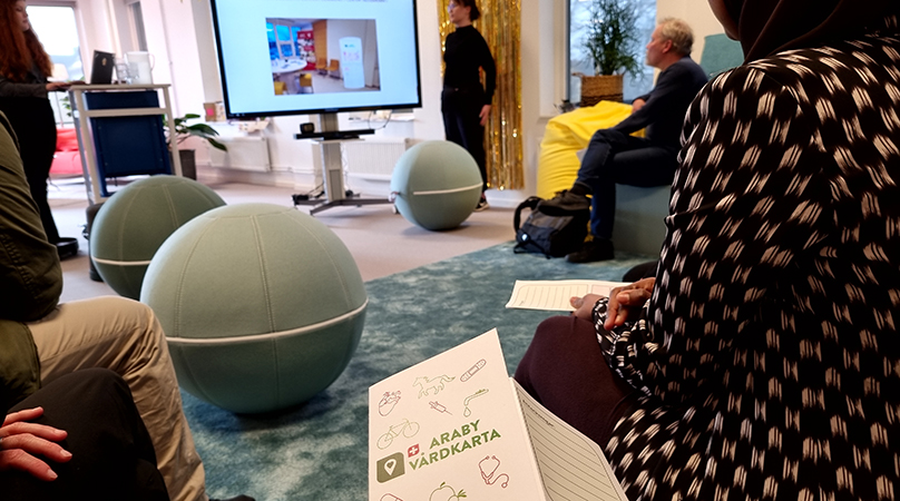 En person står framme vid en tv som visar en bild. Flera åhöra sitter och tittar mot personen och tv:n. I förgrunden syns en folder med texten "Araby vårdkarta".