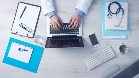 En persons händer som knappar på en laptop. Bredvid datorn ligger anteckningsblock och ett stetoskop.