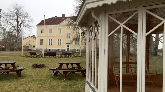 Grimslövs folkhögskola. Ett stort gult trähus med en gräsmatta, bänkar och ett lusthus framför.